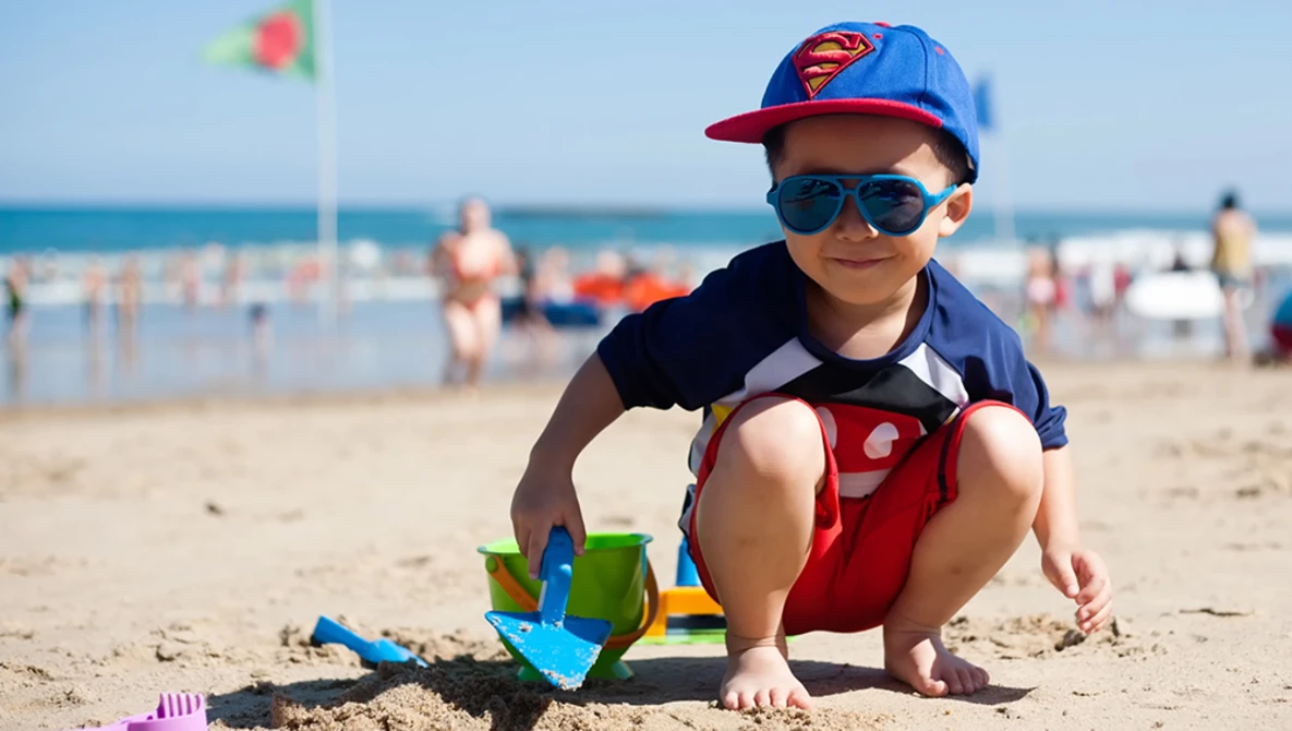 Young child crouching on sand at beach, wearing Superman-themed cap and sunglasses, playing with colorful toy beach equipment.