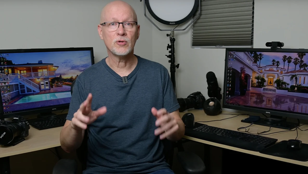Man in glasses and blue shirt gesturing while speaking in a studio with multiple monitors displaying architectural renderings.