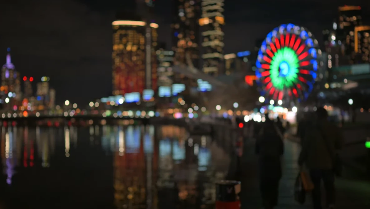 Illuminated Ferris wheel reflected in still water at night with blurred city skyline behind it.