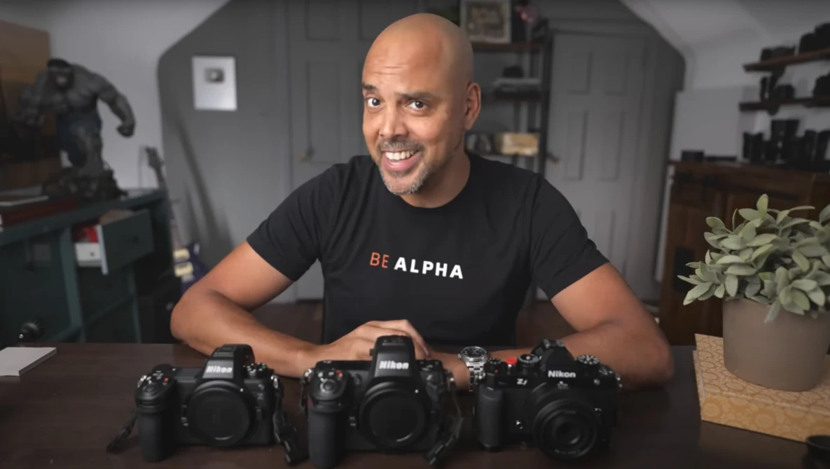 Man in black 'BE ALPHA' shirt displays three professional DSLR cameras on a desk.