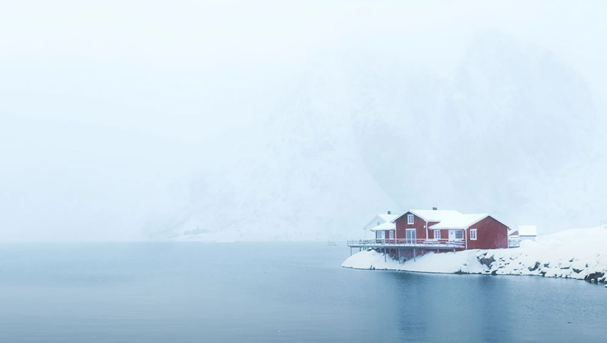 Red houses on a snowy peninsula surrounded by still, misty water in winter conditions.