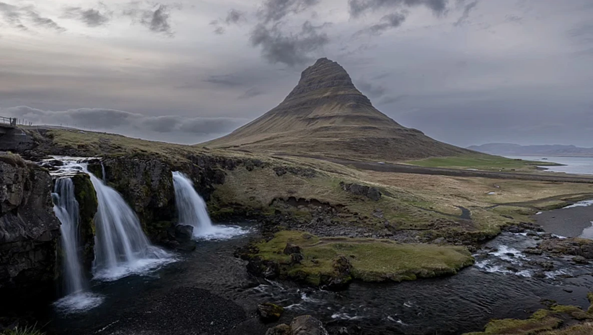 Cone-shaped mountain with cascading waterfalls in foreground under dramatic cloudy sky.