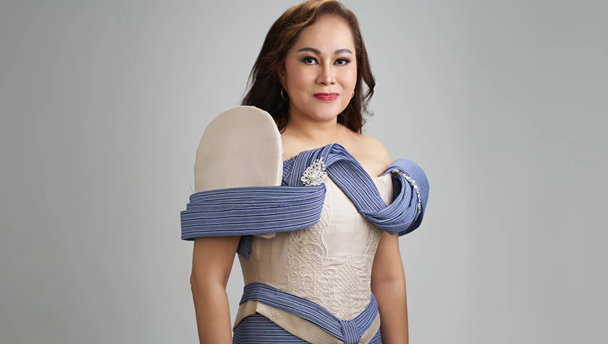 Woman in cream-colored off-shoulder dress with blue striped fabric draped around shoulders against neutral background.