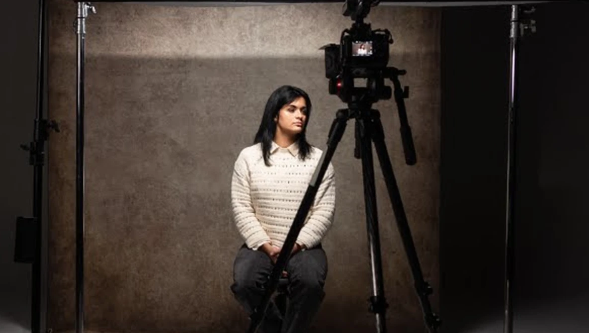 Woman in cream knit sweater seated in studio with camera mounted on tripod and professional lighting setup.