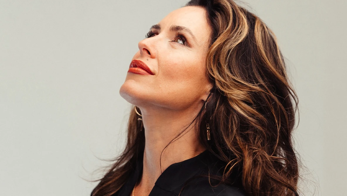 Woman with wavy brunette hair and red lipstick gazing upward against neutral background.