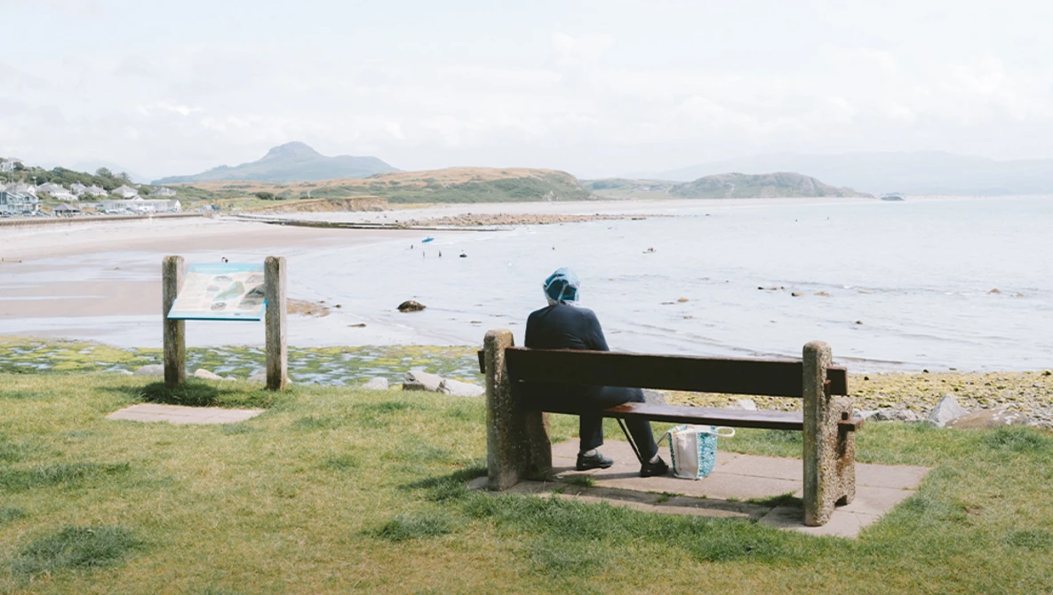 Person sitting on wooden bench overlooking a coastal beach with mountains in the distance.