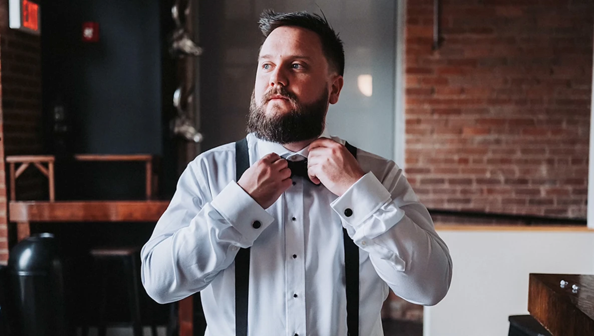 Bearded man in white formal shirt adjusting black bow tie in industrial loft setting.