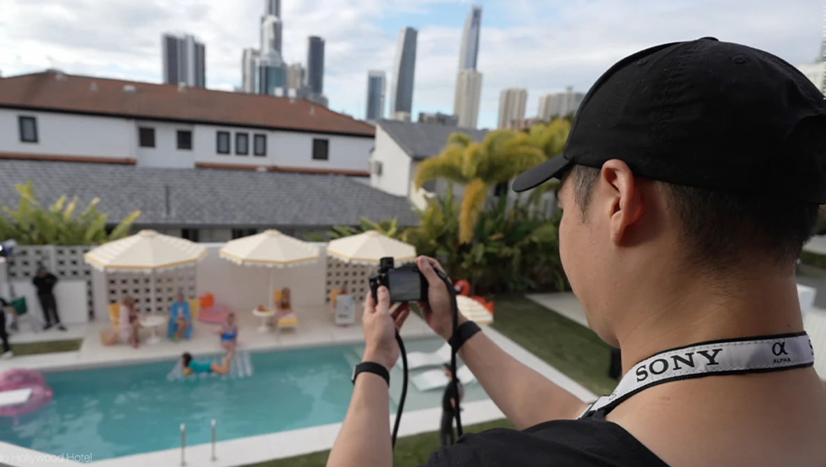 Photographer holding mirrorless camera with Sony strap, shooting poolside scene with city skyline beyond.