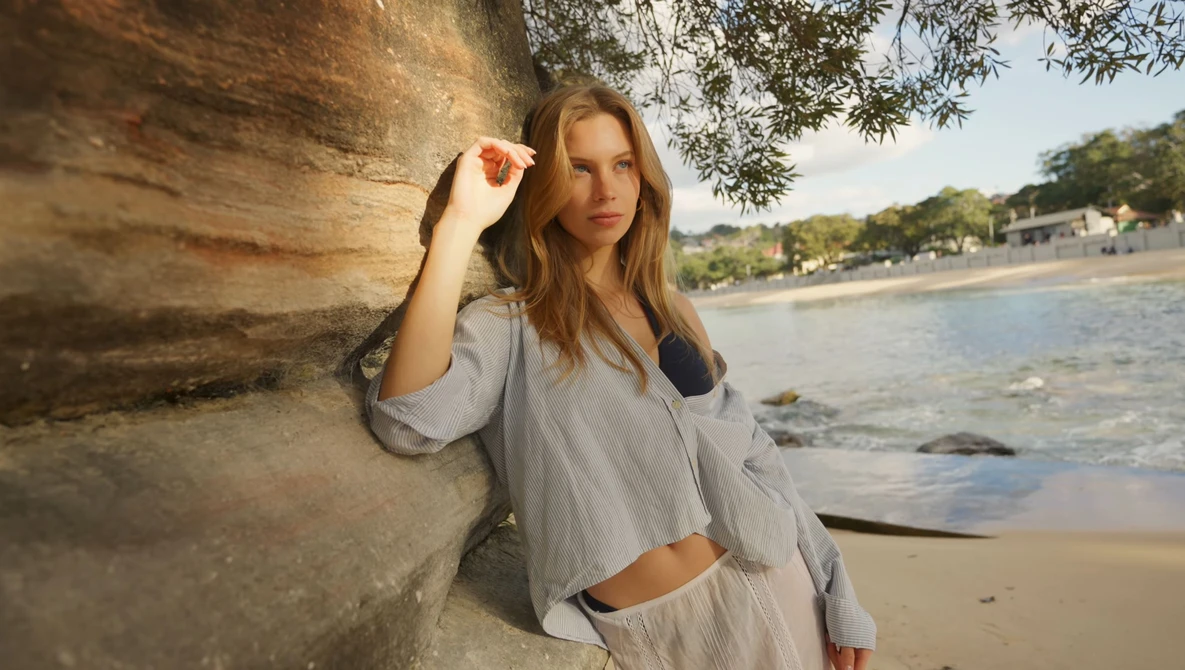 Woman in light blue shirt leaning against weathered wooden post on beach at golden hour.