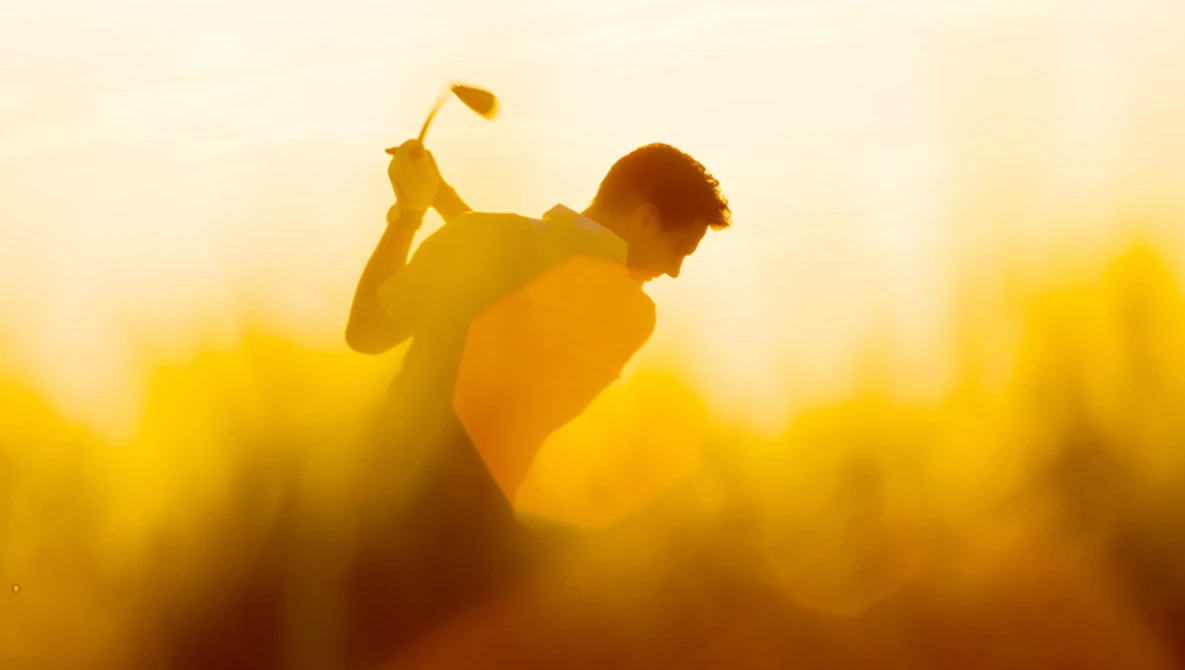 Golfer silhouetted mid-swing against golden sunset sky with blurred background.