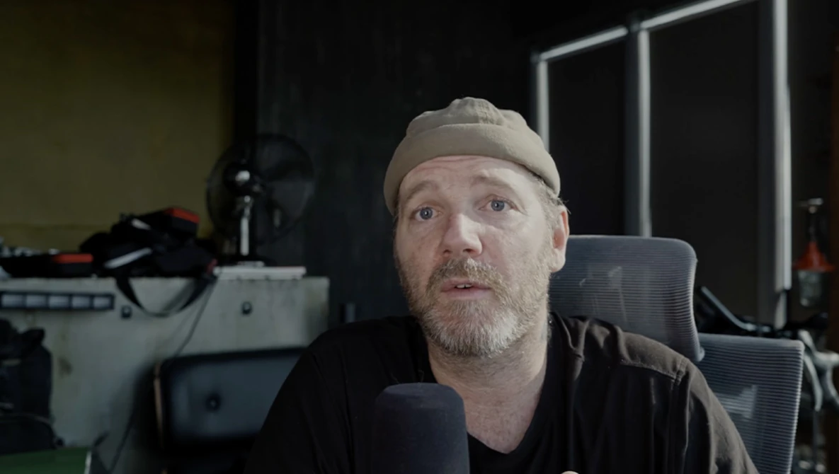 Man wearing a beanie and dark shirt speaking into a microphone in a studio setting.
