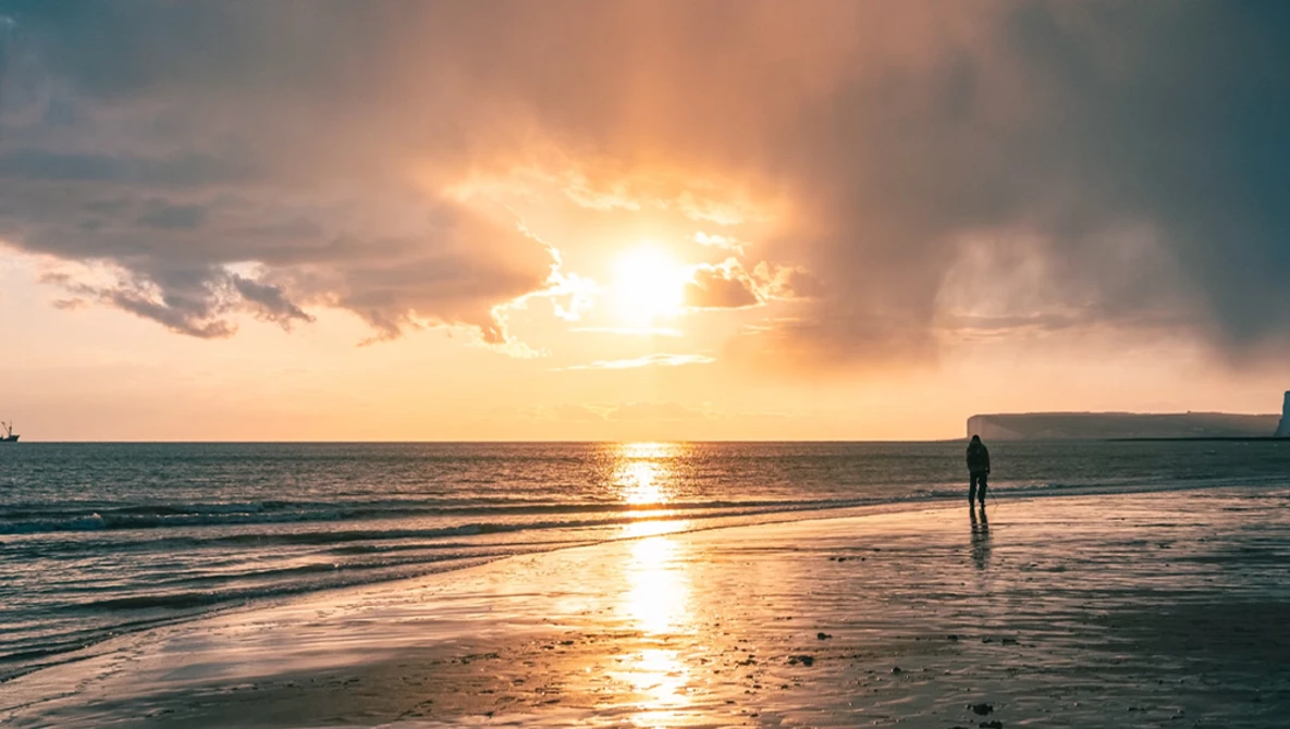 Surfer standing in shallow water during golden hour sunset with dramatic clouds and bright sun breaking through.
