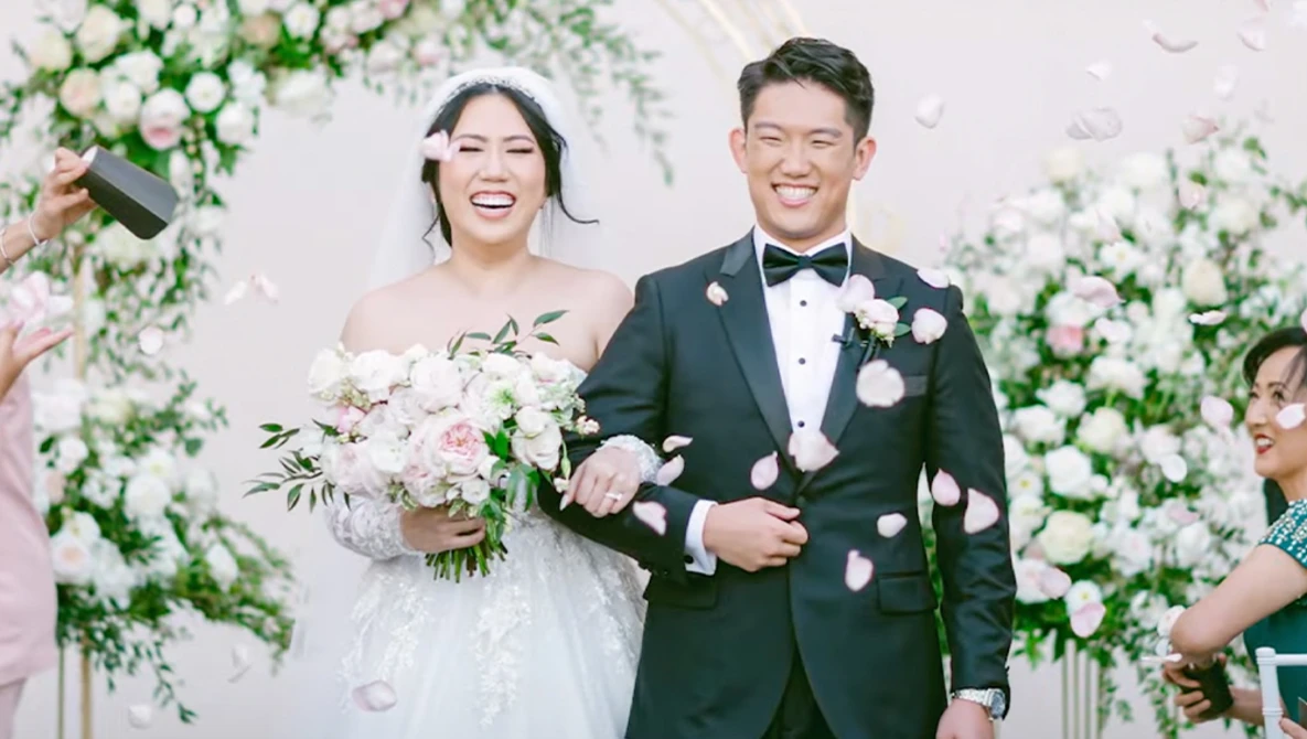 Bride and groom smiling at wedding ceremony with floral backdrop and petal confetti.