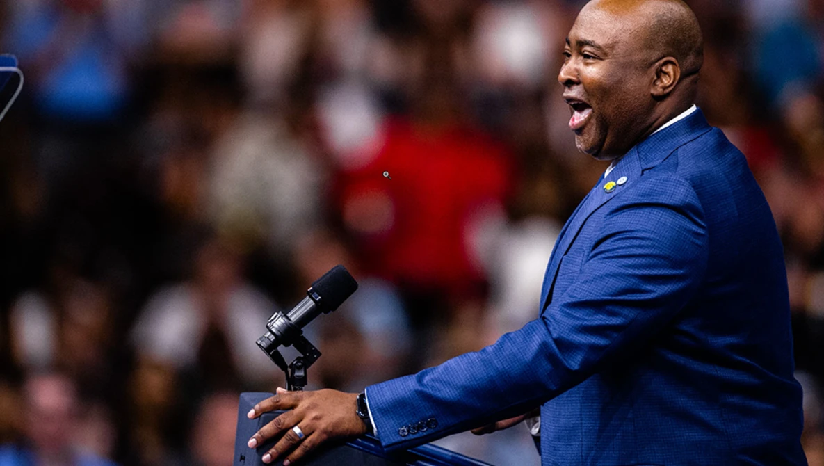 Man in blue blazer speaking at microphone before a crowd.