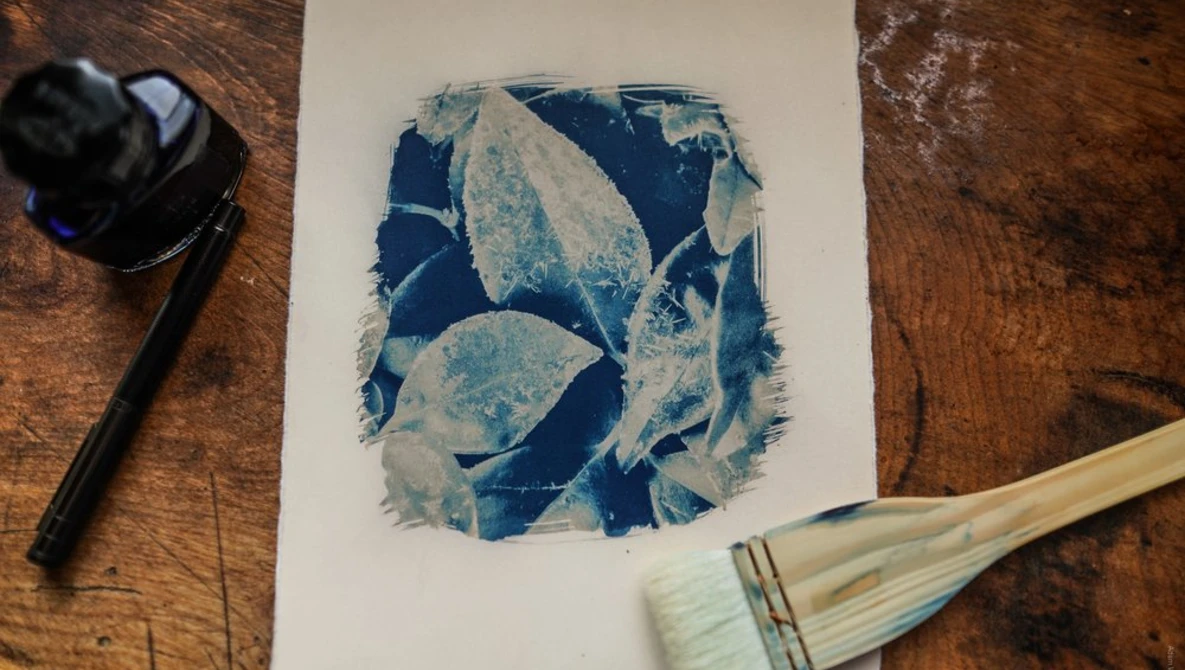 Cyanotype print with blue botanical leaf shapes on white paper, surrounded by art materials on wood surface.