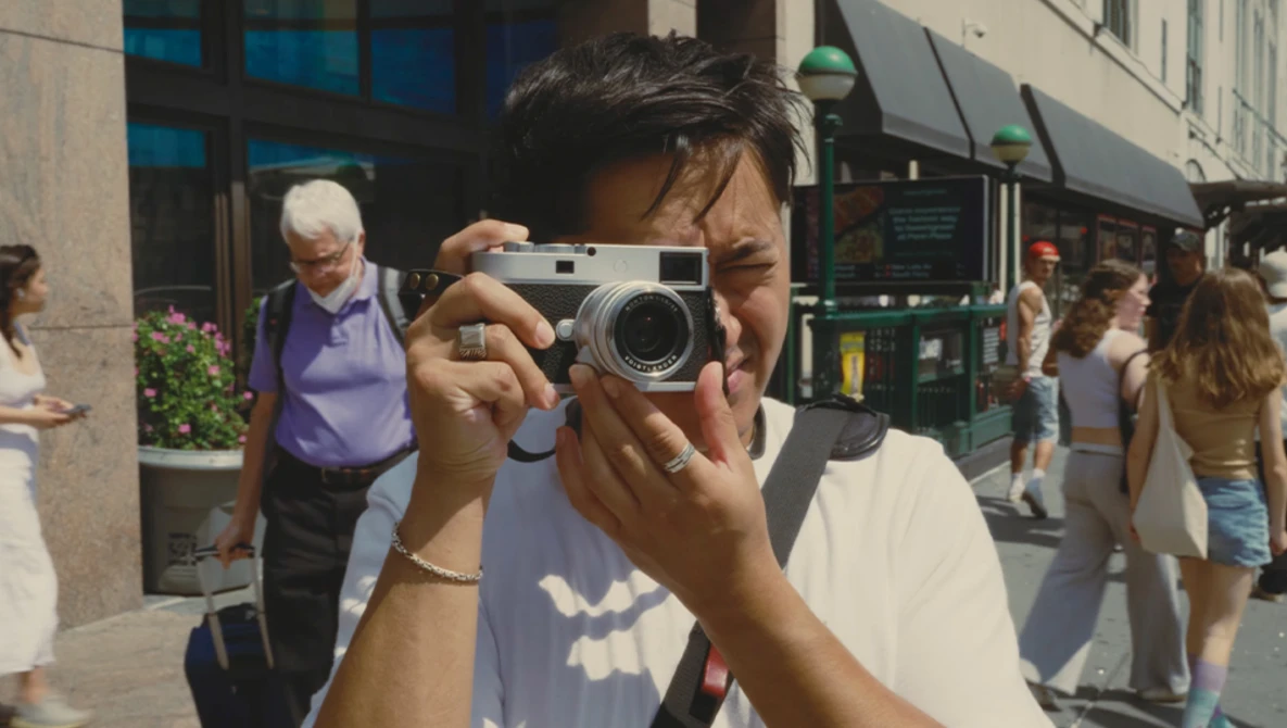 Photographer holding a rangefinder camera to their face on a sunny urban street.