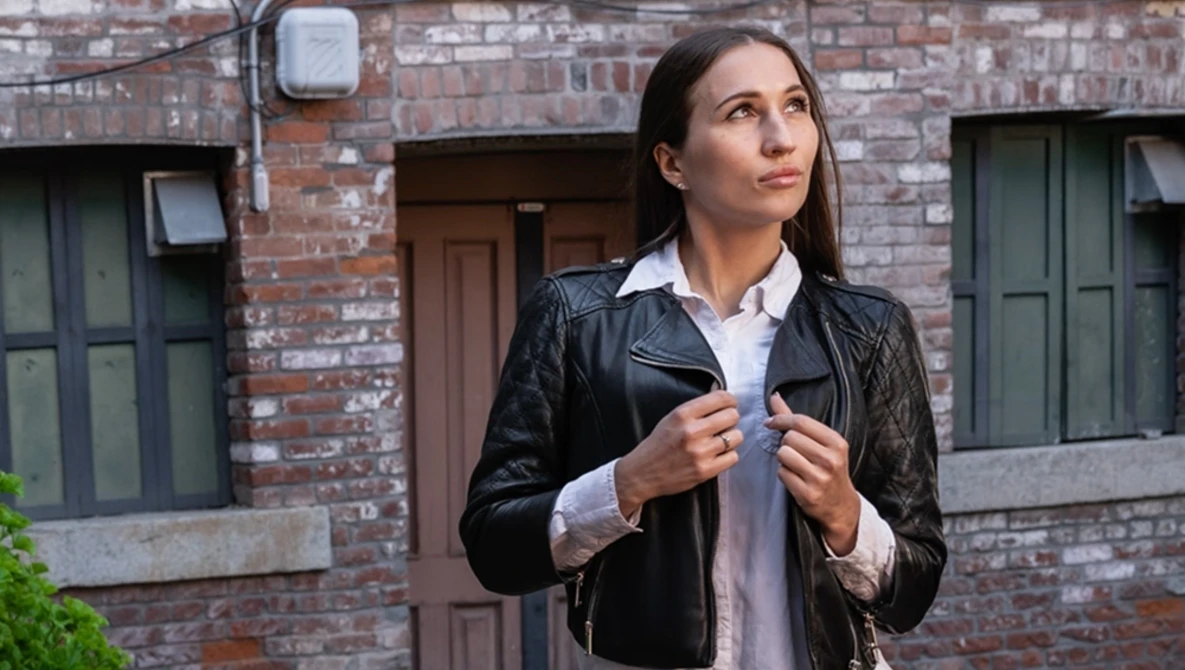 Woman in black leather jacket and white collared shirt standing against brick building backdrop.
