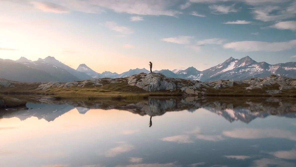 Solitary hiker standing on a rocky outcrop, reflected in still water with snow-capped mountains in the background.