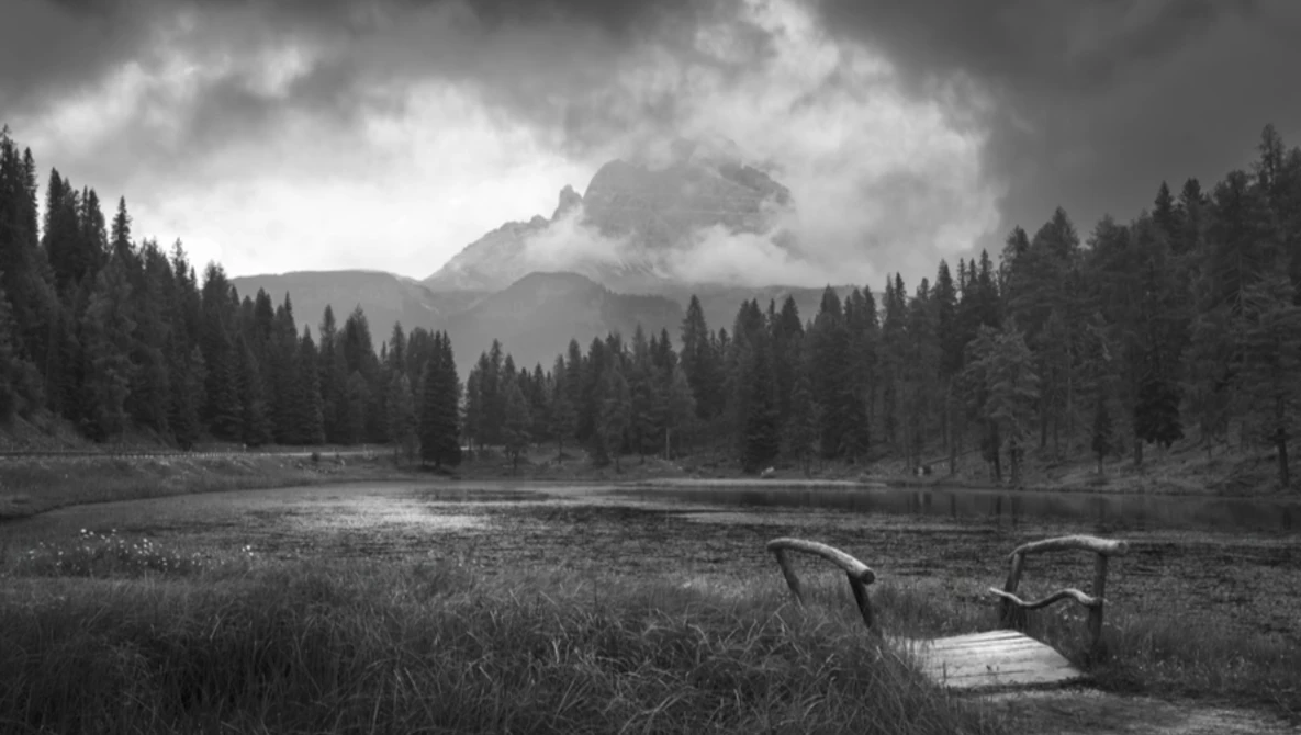Wooden dock extending into a lake surrounded by evergreen forest with a dramatic mountain peak shrouded in clouds.