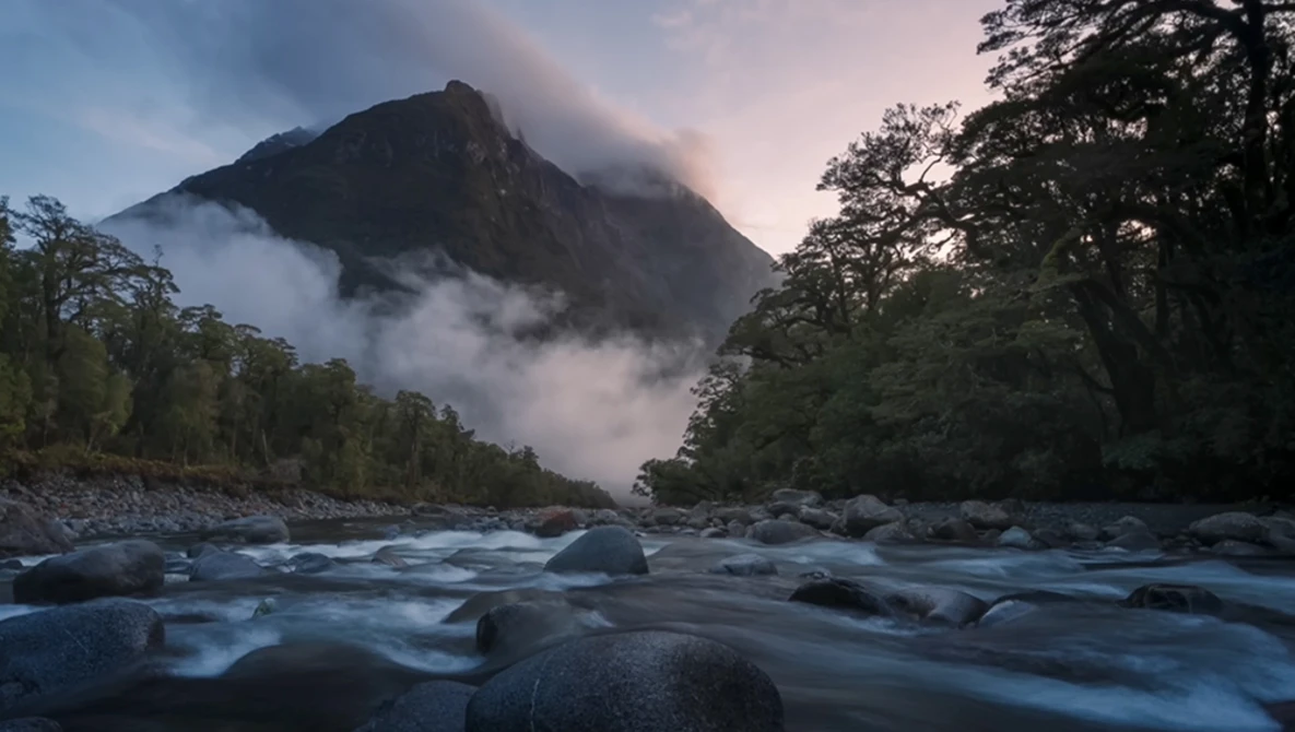 Mountain peak shrouded in mist rising above a flowing river with forested banks at twilight.