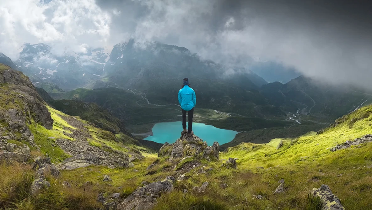 Hiker in blue jacket standing on rocky outcrop overlooking turquoise alpine lake surrounded by misty mountains.