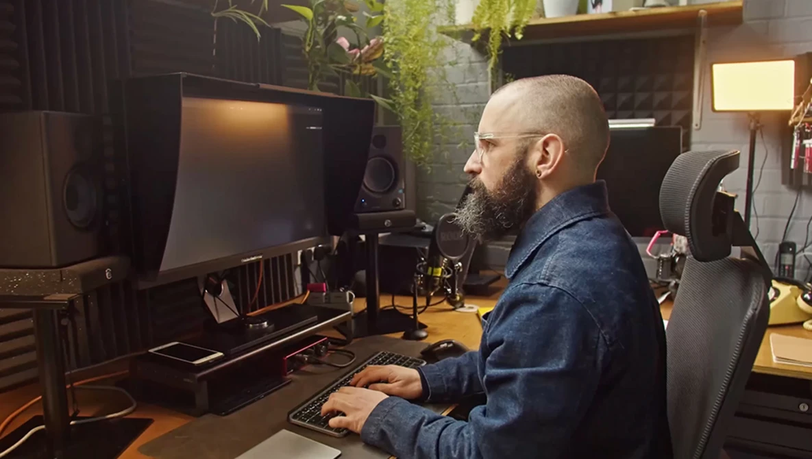 Bearded man working at a computer desk in a professional studio setup with monitor, keyboard, and audio equipment.
