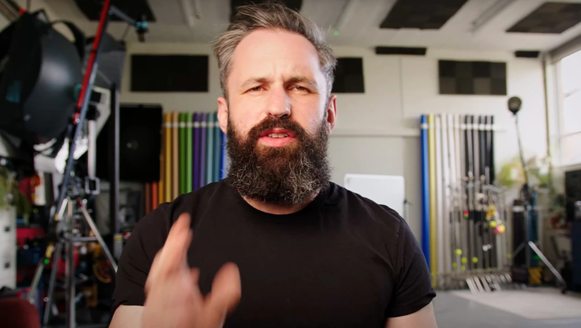 Bearded man in black t-shirt gesturing while speaking in a studio with professional lighting equipment visible.