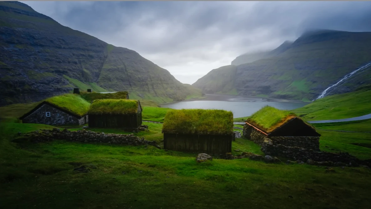 Turf-roofed stone cottages nestled in a verdant valley with mountains, a glacial lake, and a distant waterfall.
