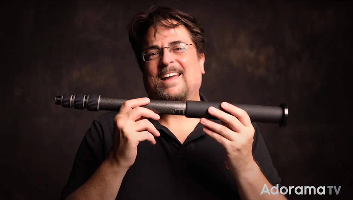 Man wearing glasses holding a telephoto lens toward the camera against a dark background.