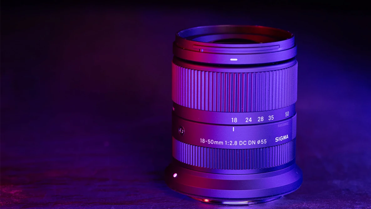 Sigma 18-50mm f/2.8 DC DN lens illuminated with purple and magenta neon lighting against a dark background.