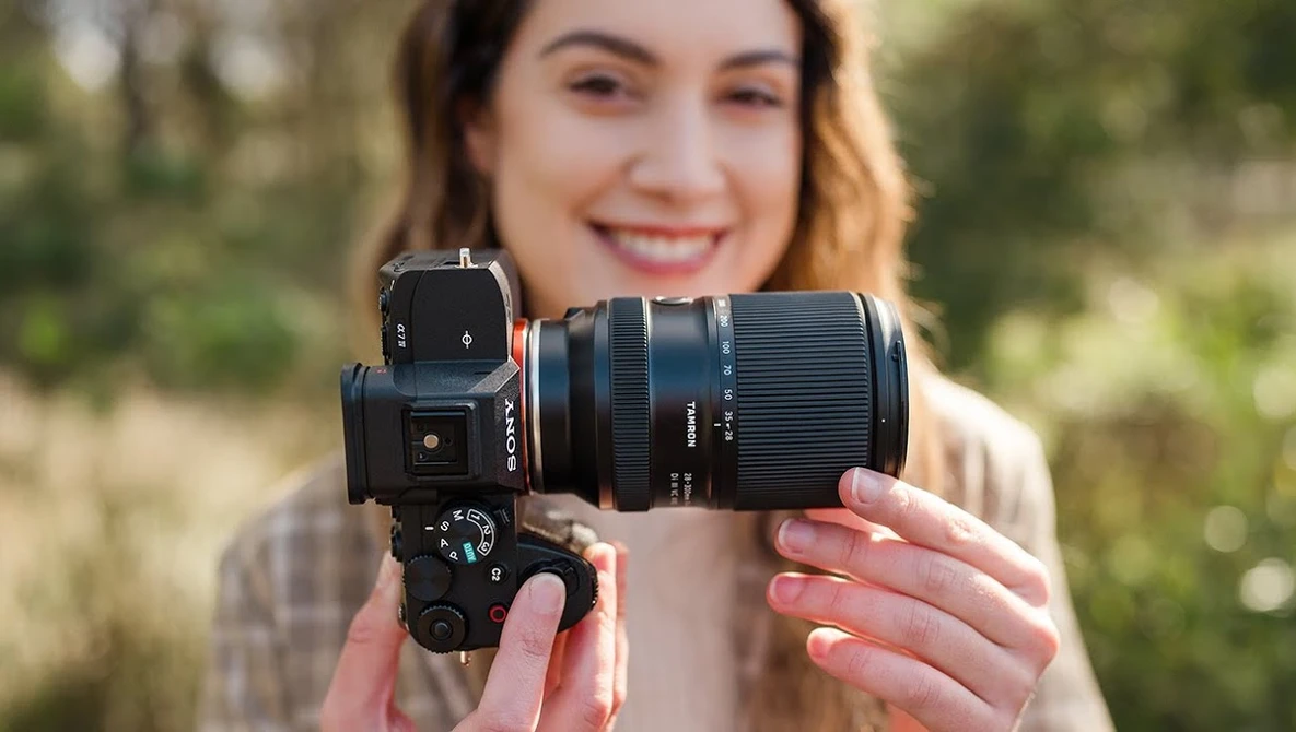 A photographer holds a mirrorless camera with an attached telephoto lens toward the camera, smiling outdoors.
