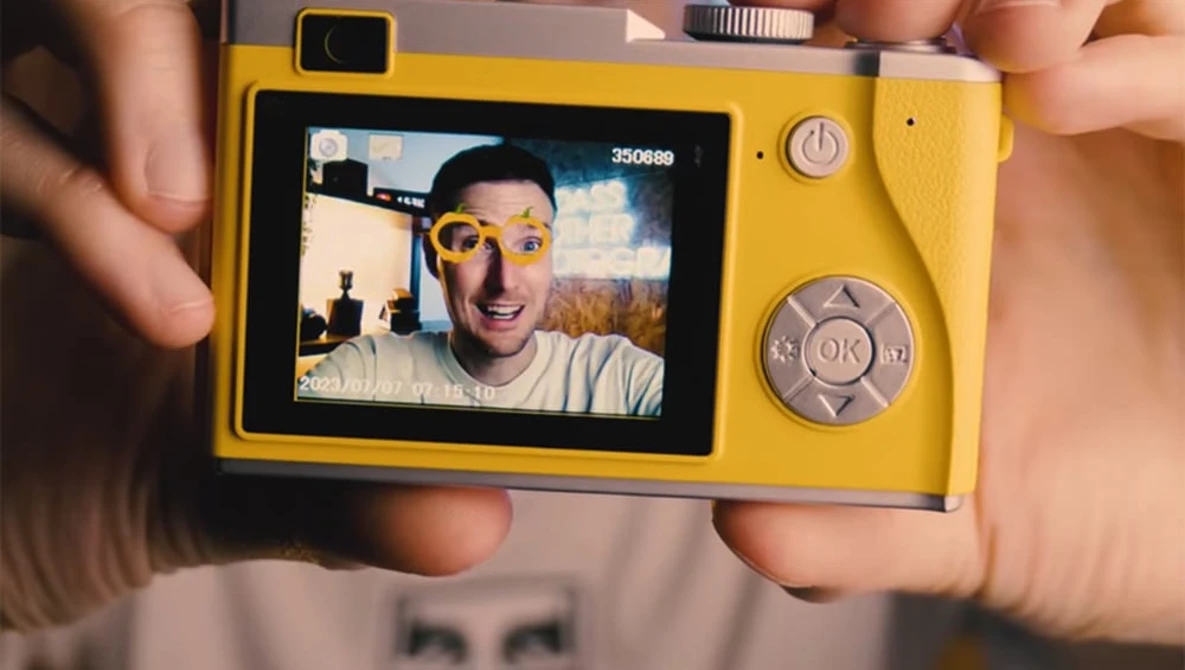 Yellow compact digital camera displaying a selfie of a person wearing yellow-tinted sunglasses on its rear LCD screen.