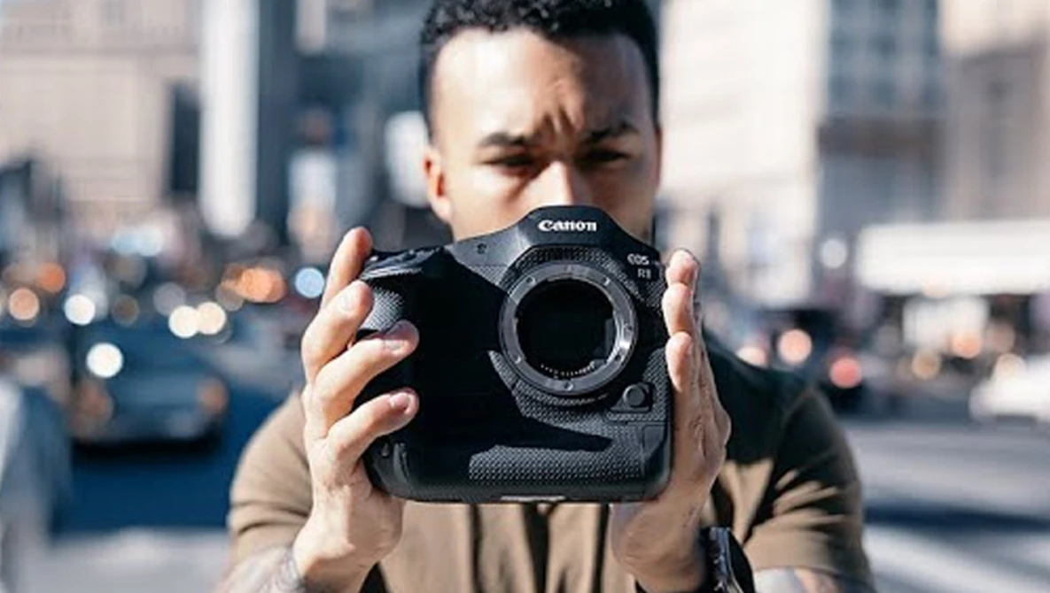 Photographer holding Canon EOS R5 camera body in urban setting with blurred cityscape background.