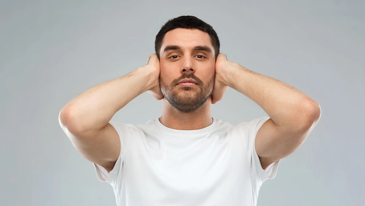 Man in white t-shirt with hands behind head against neutral gray background.