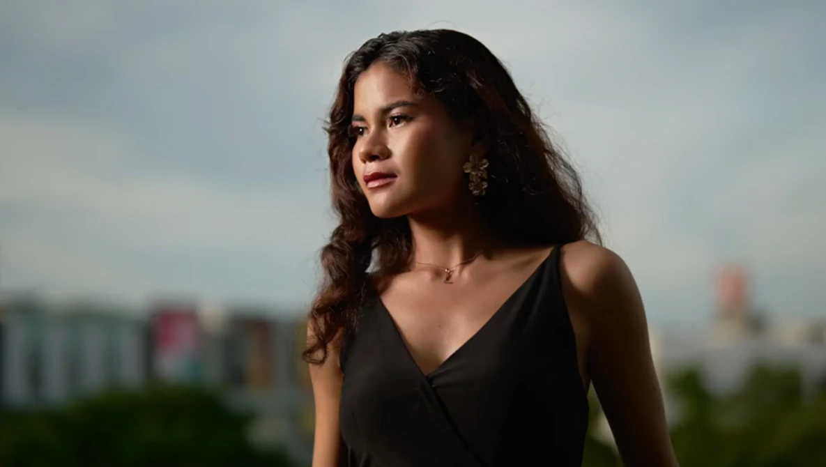 Woman with long dark wavy hair wearing black sleeveless top and gold earrings, photographed outdoors against blurred cityscape.