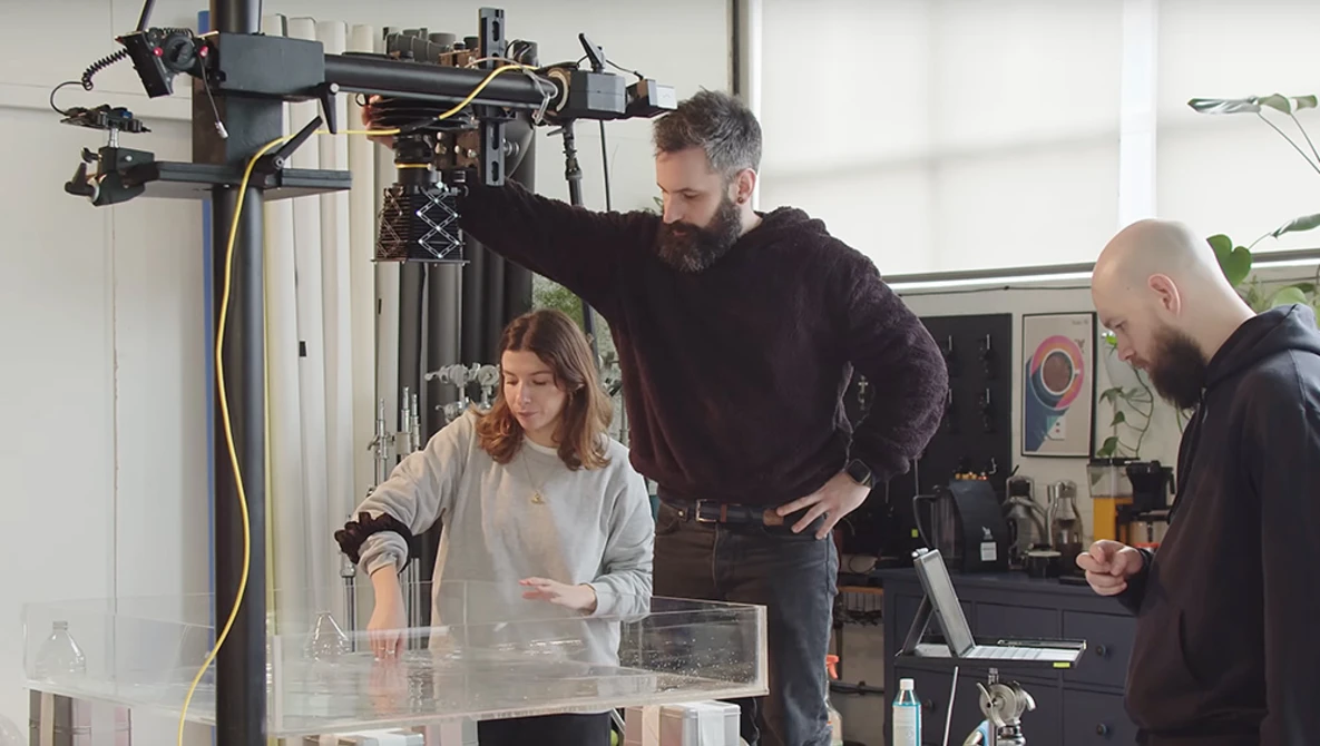 Three people collaborating around a 3D printer in a bright workshop studio setting.