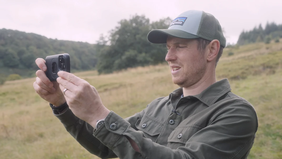 Man in cap holding smartphone to capture landscape photo in rolling hills.