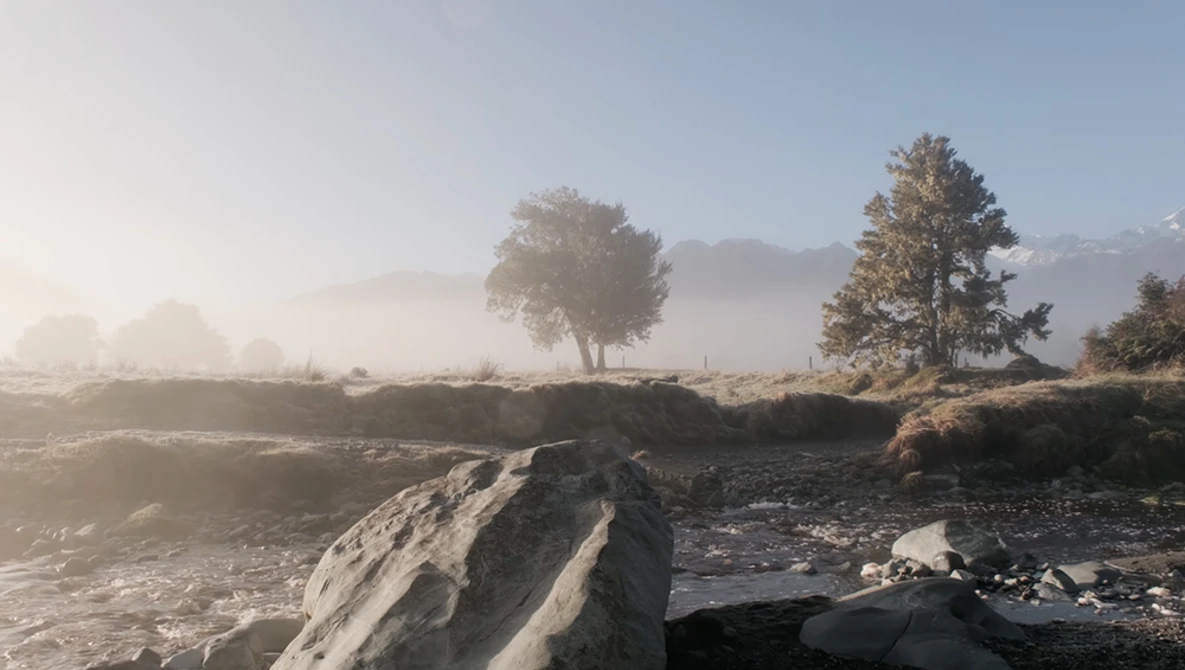 Misty landscape with trees and large boulders in foreground along a stream.