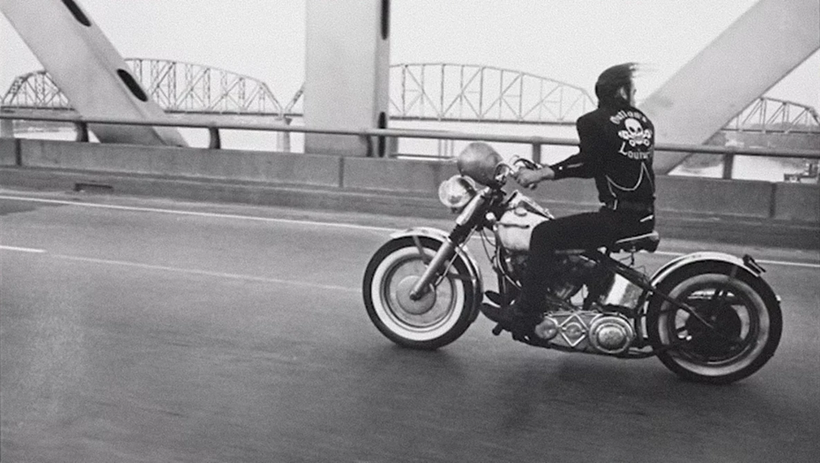 Motorcyclist riding a cruiser on a bridge with architectural steel framework visible in the background.