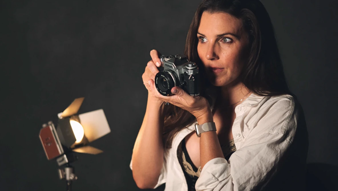 Female photographer holding a vintage SLR camera, illuminated by studio lighting against a dark background.