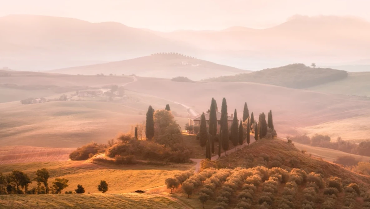 Tuscan villa surrounded by cypress trees on a hilltop in misty golden landscape.