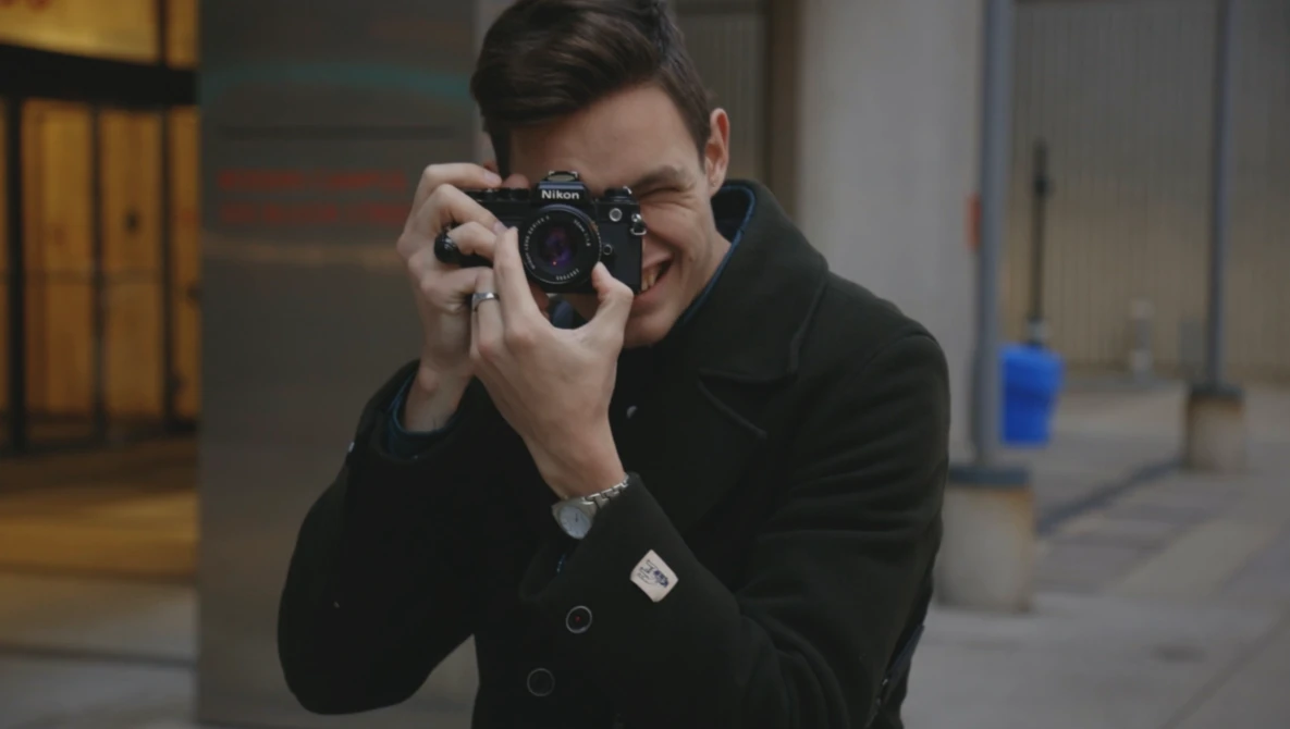 Young man in black jacket holding a rangefinder camera to his face, smiling while shooting in an urban corridor.