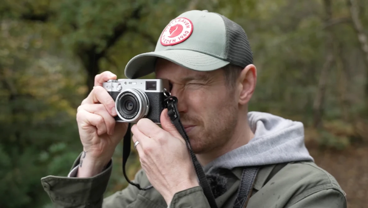 Photographer holding a rangefinder camera up to his eye while wearing a cap and olive jacket.