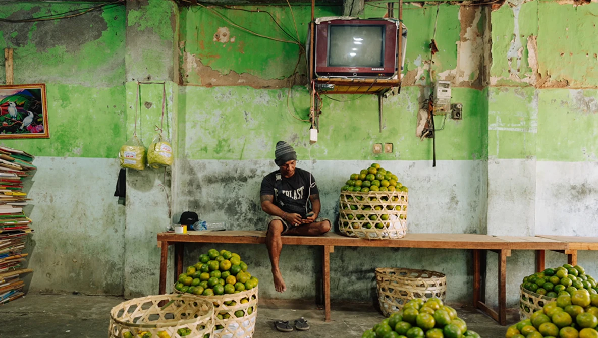 Man sitting on wooden bench in front of green deteriorated wall surrounded by baskets of limes.