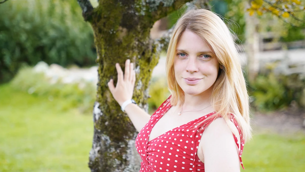 Young woman with blonde hair wearing a red polka dot dress, leaning against a moss-covered tree in a garden setting.