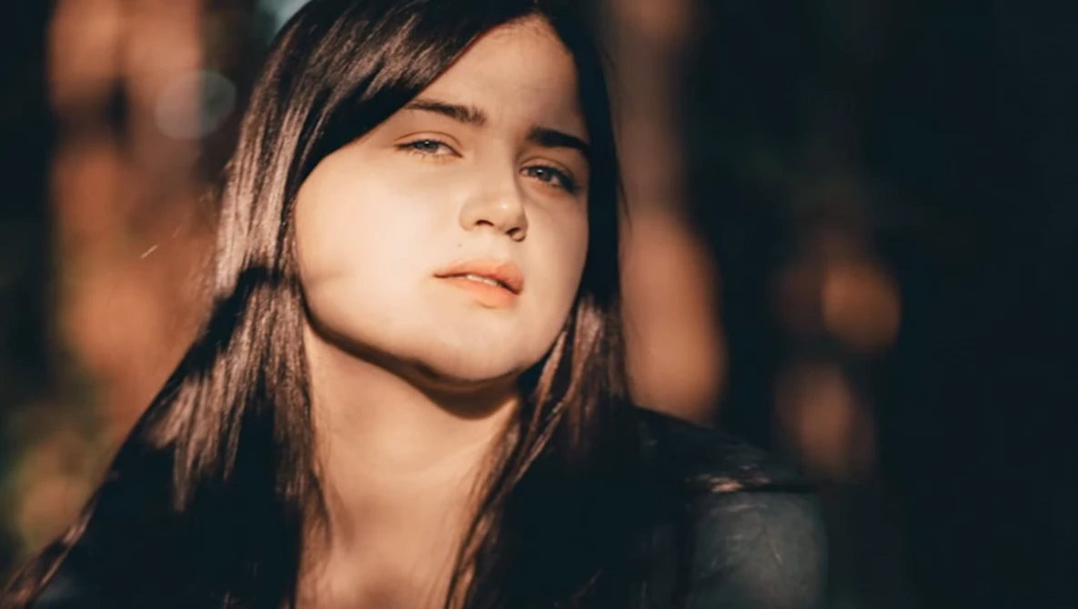 Young woman with long dark hair gazing upward in warm golden light against a dark background.