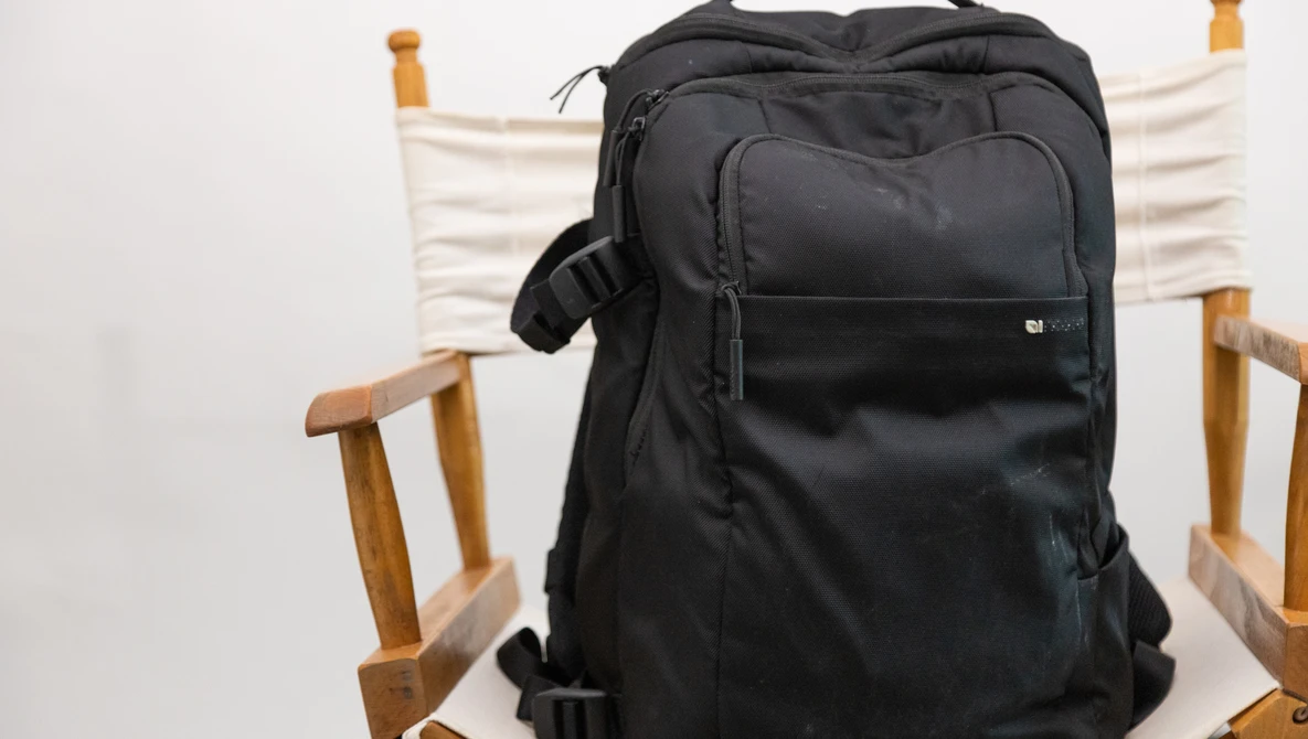 Black camera backpack displayed on a wooden chair with white canvas seat and back.
