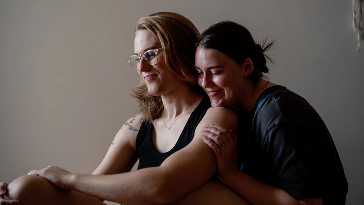 Two smiling people sitting together against a neutral gray background, looking downward in a candid moment.