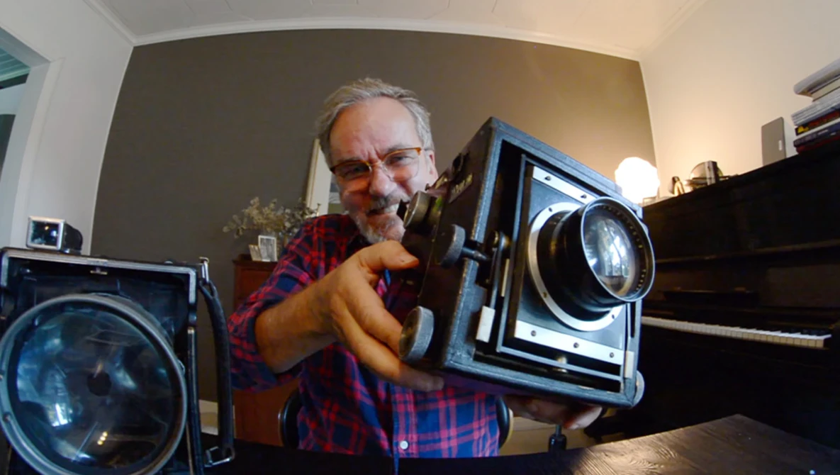 Photographer demonstrating a large format film camera held up to his face in an indoor studio setting.