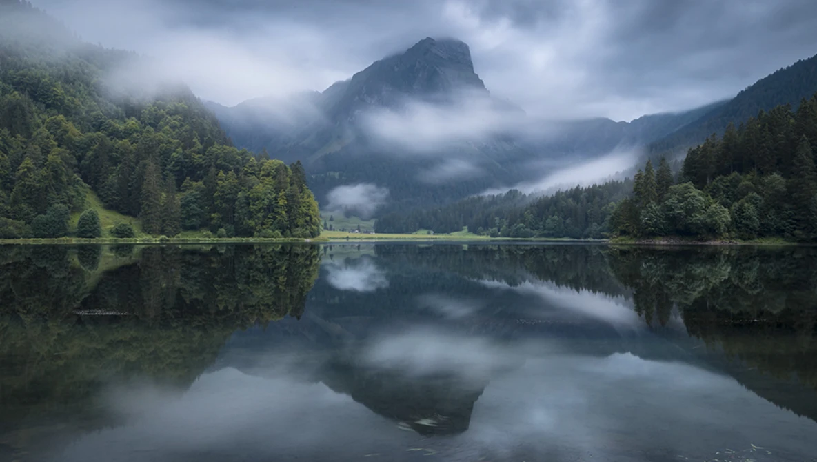 Mountain lake with forested shores and misty peaks reflected in calm water.